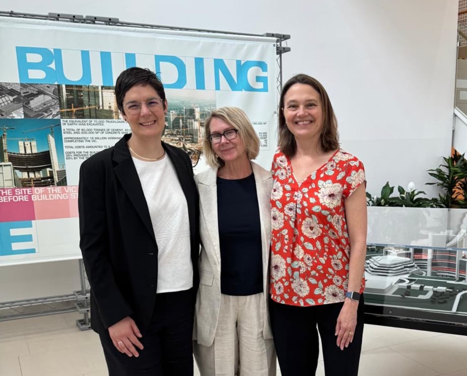 Three women stand together smiling in front of a display titled "BUILDING." The woman on the left wears a black suit with a white top and glasses, the woman in the center wears a light-colored suit and glasses, and the woman on the right wears a red floral blouse and black pants. The setting is an indoor exhibition or academic space with informational posters and a model structure in the background.